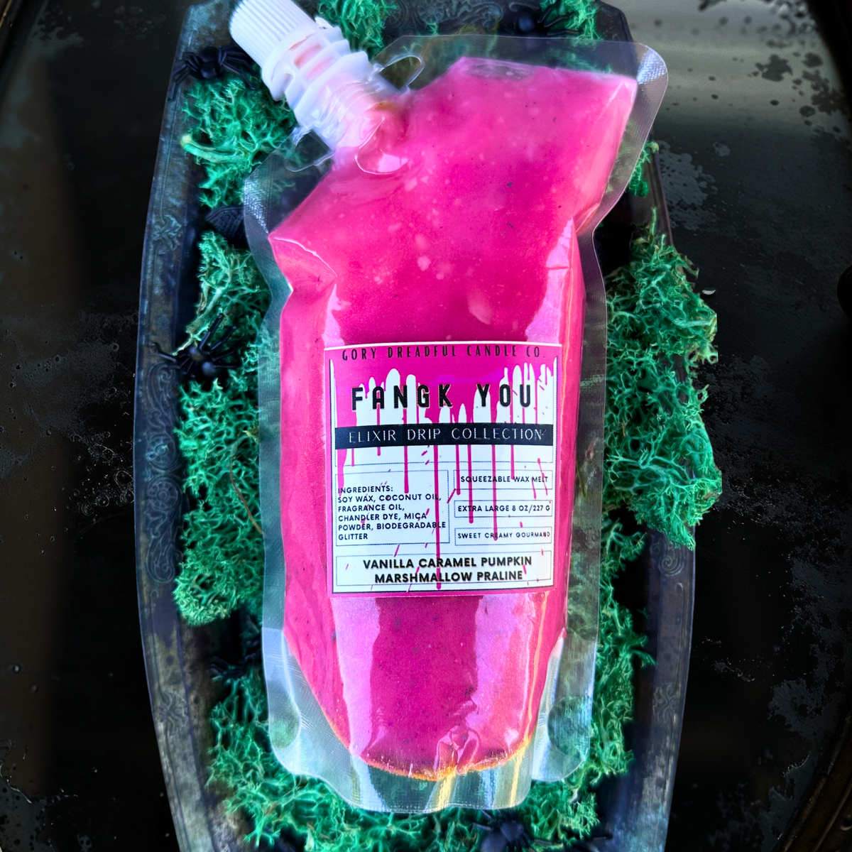 A clear squeeze pouch filled with bright pink liquid, labeled 'Frank You Luxur Drip Collection' and 'Vanilla Caramel Pumpkin Marshmallow Franchise,' resting on green moss inside a dark tray.