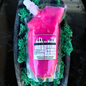 A clear squeeze pouch filled with bright pink liquid, labeled 'Frank You Luxur Drip Collection' and 'Vanilla Caramel Pumpkin Marshmallow Franchise,' resting on green moss inside a dark tray.